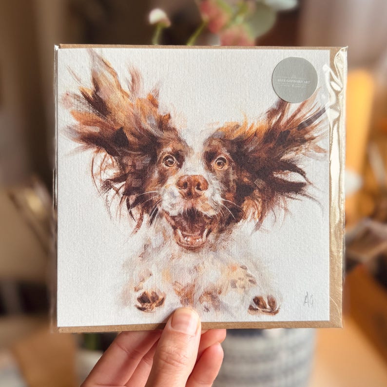 Op de afbeelding: Een vierkante wenskaart met een aquarel schilderij van een hond met bruin en wit haar, zijn oren wapperen. De hond heeft een open mond en lijkt te glimlachen. De kaart zit op een bruine achtergrond en heeft een kleine, ronde sticker.