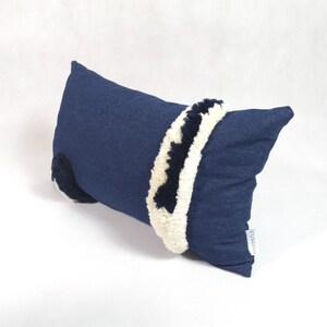May include: A blue denim pillow with white and black pom-poms around the edges. The pillow has a white label with the word "cushions" on it.