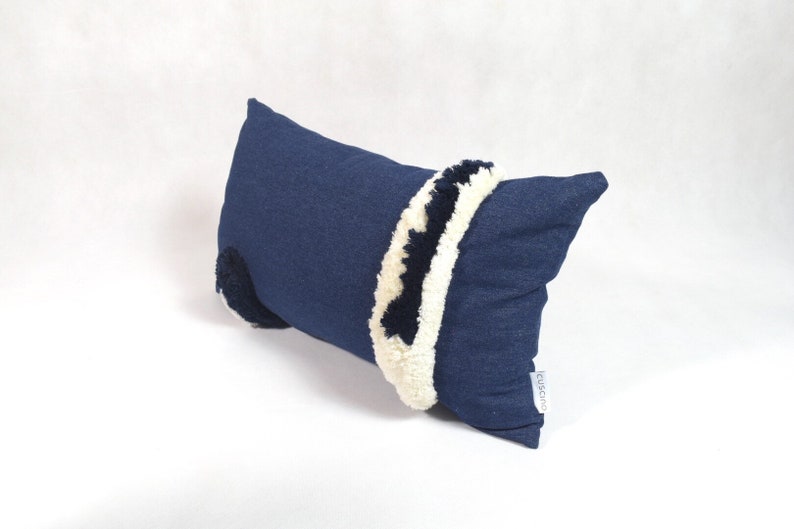 May include: A blue denim pillow with white and black pom-poms around the edges. The pillow has a white label with the word "cushions" on it.