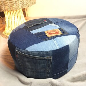 Op de afbeelding: Ronde denim poef gemaakt van hergebruikte jeans. Het patchwork ontwerp toont verschillende tinten blauwe denim, inclusief zakken en het Levi Strauss & Co. 508 label. Een uniek en stijlvol stuk voor elke woonruimte.