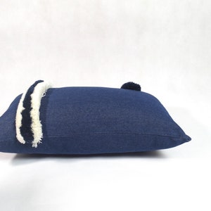 May include: A blue denim pillow with white and black trim and a pom-pom on one end.