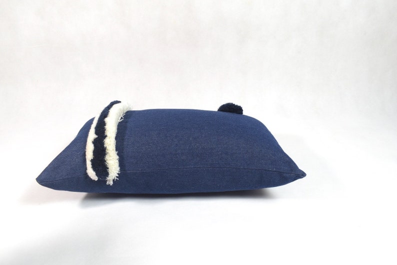 May include: A blue denim pillow with white and black trim and a pom-pom on one end.