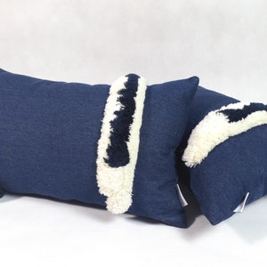 May include: Two blue denim throw pillows with white and black fringe trim. The pillows are rectangular and have a soft, textured surface.