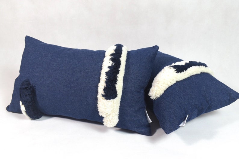 May include: Two blue denim throw pillows with white and black fringe trim. The pillows are rectangular and have a soft, textured surface.