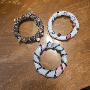 May include: Three handmade fabric rings with decorative beads and pins. One ring is wrapped in a brown and black camouflage pattern. Another is wrapped in white fabric with colorful beads. The third ring is wrapped in white fabric with blue beads.