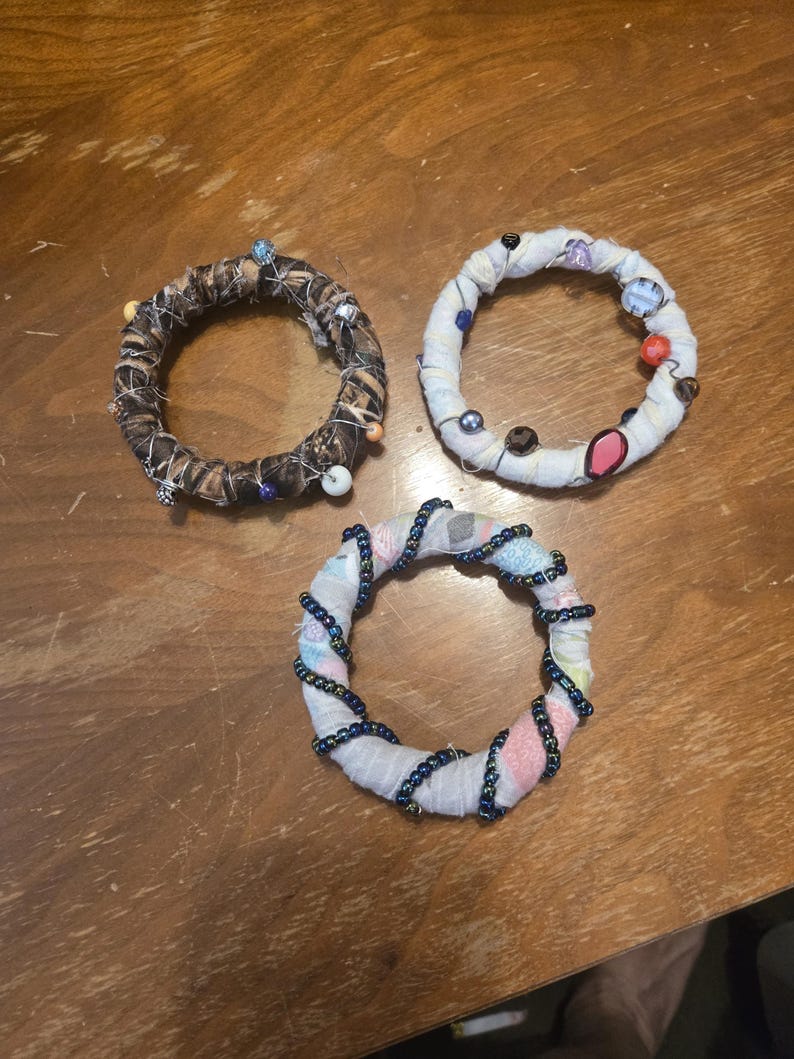 Puede incluir: Tres anillos de tela hechos a mano con cuentas y alfileres decorativos. Un anillo est&aacute; envuelto en un patr&oacute;n de camuflaje marr&oacute;n y negro. Otro est&aacute; envuelto en tela blanca con cuentas de colores. El tercer anillo est&aacute; envuelto en tela blanca con cuentas azules.