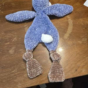 K&ouml;nnte beinhalten: Ein handgefertigtes Pl&uuml;sch-Hasenspielzeug in Blau- und Braunt&ouml;nen. Der Hase hat lange Ohren, einen wei&szlig;en Schwanz und braune F&uuml;&szlig;e. Das weiche, strukturierte Material verleiht ihm ein kuscheliges Aussehen.
