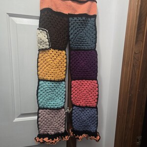May include: Handmade crochet pants in a patchwork design. The pants feature a variety of colors, including orange, black, brown, yellow, teal, purple, pink, and blue. The pants have a ruffled hem and are displayed on a hanger.