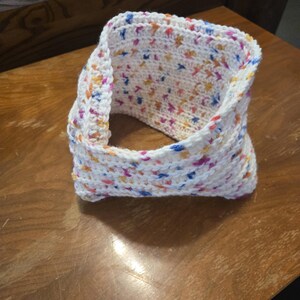 May include: A white crocheted cowl with colorful flecks of blue, pink, orange, and yellow. The handmade neck warmer is a tube shape, folded over on itself. The cowl is sitting on a wooden surface.