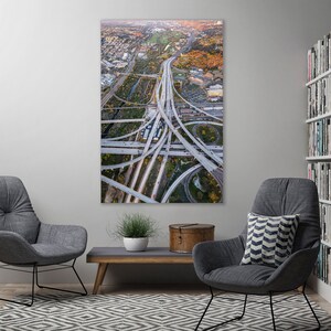 May include: An aerial view photograph of a complex highway interchange with numerous overpasses and ramps. The image captures the intricate network of roads, with cars and trucks visible. The surrounding area includes buildings and trees, with a warm, autumnal color palette.