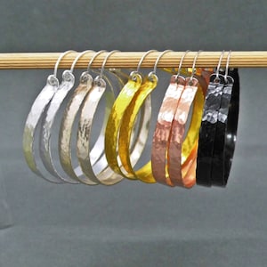 Könnte beinhalten: Ein Set aus sechs gehämmerten Creolen in verschiedenen Metallic-Oberflächen, darunter Silber, Gold, Kupfer und Schwarz. Die Ohrringe werden auf einem Holzstab präsentiert.