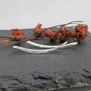 Könnte beinhalten: Ein Paar silberne, gehämmerte Ohrclips, gebogen und leicht verbogen, auf einer dunkelgrauen Schieferfläche.