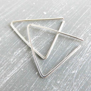 May include: Two silver-coloured, triangular earrings. The earrings have a simple, geometric design. They are displayed on a grey, textured surface. The earrings are a minimalist style.