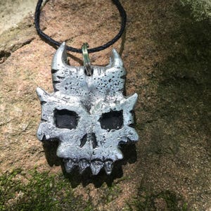 May include: A silver skull pendant with horns, detailed with textured ridges and black eye sockets, hanging from a black cord.