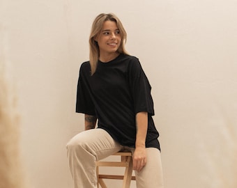 May include: A person wearing a black t-shirt and beige pants, sitting on a wooden step stool. The person is also wearing white boots. The background is a light beige color.