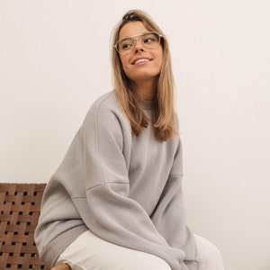 Puede incluir: Una sudadera gris claro y pantalones blancos. La modelo lleva gafas con montura dorada y está sentada en una silla de mimbre marrón. El conjunto es informal y cómodo, adecuado para el uso diario.