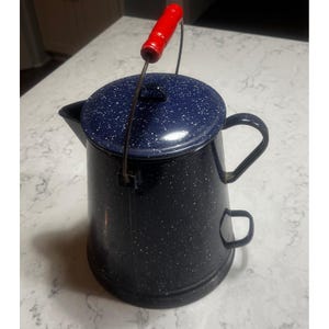 May include: A black enamel coffee pot with a speckled navy blue lid and a red handle. The pot has a spout and two handles, one on the side and one for the lid. The pot is designed for camping or outdoor use.