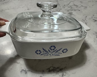 May include: A white casserole dish with a clear glass lid and handle. The dish features a blue floral design on the front. The lid has a clear glass knob. The dish is on a white surface.