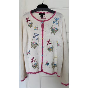 May include: Cream-colored cardigan sweater with embroidered basket and floral designs. Features a pink trim along the neckline, button placket, and cuffs. The sweater has a button-up front and long sleeves.