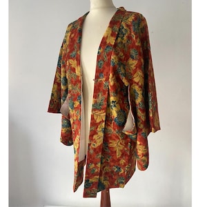 Könnte beinhalten: Ein offener Kimono in leuchtenden Farben mit einem botanischen Muster aus Blättern und Blumen. Die dominierenden Farben sind Rot, Gelb und Blau. Der Kimono wird auf einer Schaufensterpuppe präsentiert, die seinen fließenden Schnitt und die weiten Ärmel zeigt.