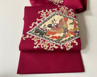 以下が含まれることがあります： 赤い絹の生地に、金と白の刺繍で鳳凰がダイヤモンド型にデザインされています。生地は半分に折り畳まれており、デザインは前面に見えます。