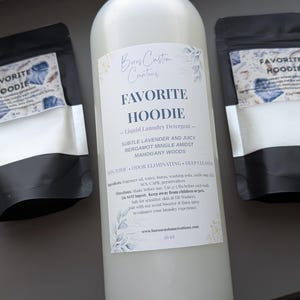 Op de afbeelding: Een grote fles Favorite Hoodie vloeibaar wasmiddel tussen twee kleinere zwarte zakjes waspoeder. De etiketten zijn voorzien van bloemmotieven en tekst die het geurprofiel van lavendel, bergamot en mahoniehout beschrijft.