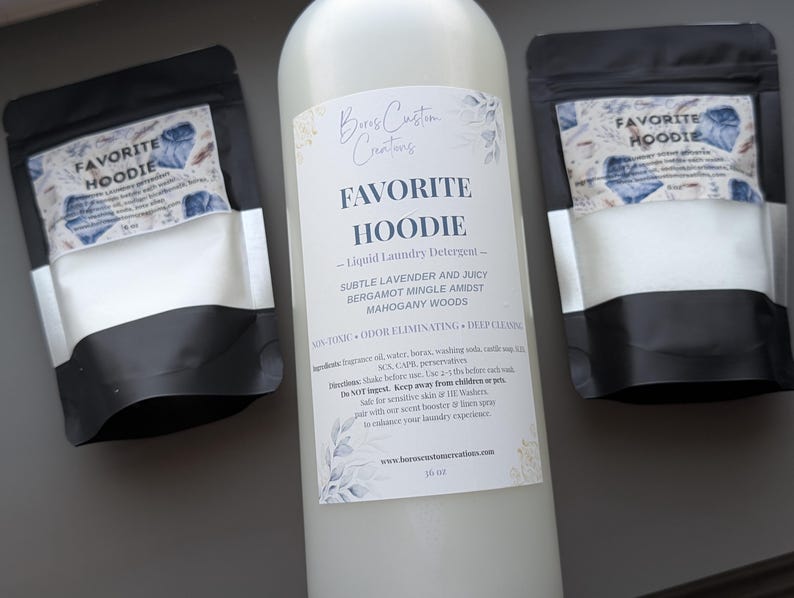 Op de afbeelding: Een grote fles Favorite Hoodie vloeibaar wasmiddel tussen twee kleinere zwarte zakjes waspoeder. De etiketten zijn voorzien van bloemmotieven en tekst die het geurprofiel van lavendel, bergamot en mahoniehout beschrijft.