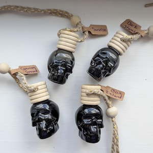 May include: Four black glass skull-shaped car air freshener bottles with light wood caps, hanging from braided beige and gold cords with wooden beads. Each bottle has a small brown paper tag labelled Sunrise, Dark Storms, Common Ground, or Oak Island.