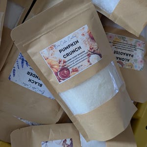 May include: A brown paper pouch containing white laundry scent booster crystals. The label features the text PUMPKIN CRUNCH, LAUNDRY SCENT BOOSTER, 10 oz, and ingredients. A clear window reveals the white granular contents inside the sealed bag.