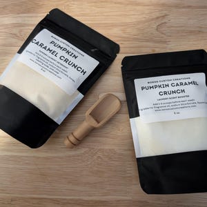 May include: Two black pouches with white labels containing laundry scent booster powder. The labels read "Pumpkin Caramel Crunch" and include the brand name "Boros Custom Creations". The pouches are on a wooden surface with a small wooden scoop.