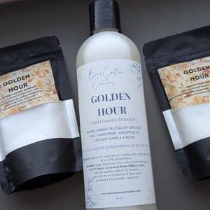 Op de afbeelding: Een fles van 473 ml Golden Hour vloeibaar wasmiddel met een zwarte dop, geflankeerd door twee zwarte zakjes van 170 g met Golden Hour wasparfum en waspoeder. Alle etiketten zijn voorzien van bloemmotieven en gedetailleerde productinformatie.