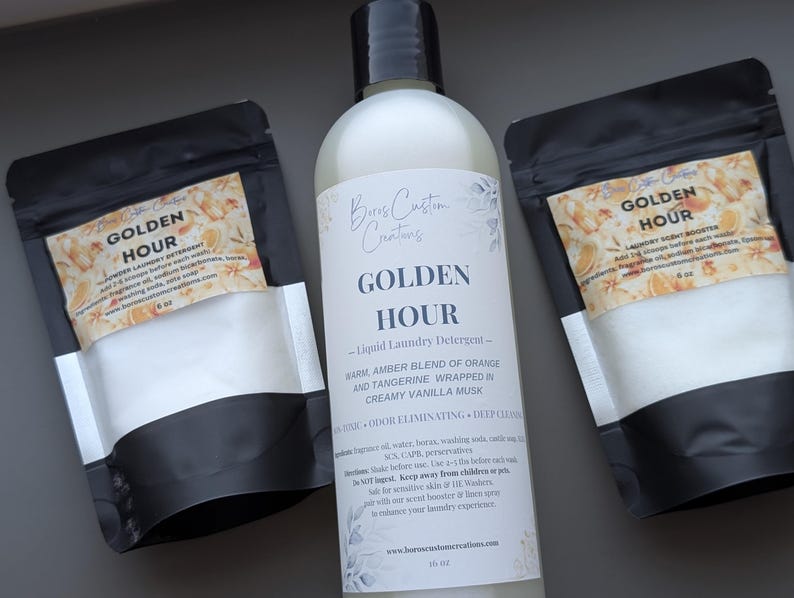 Op de afbeelding: Een fles van 473 ml Golden Hour vloeibaar wasmiddel met een zwarte dop, geflankeerd door twee zwarte zakjes van 170 g met Golden Hour wasparfum en waspoeder. Alle etiketten zijn voorzien van bloemmotieven en gedetailleerde productinformatie.