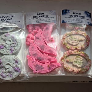 Puede incluir: Variedad de ceras perfumadas en envases transparentes. La cera "Favorite Hoodie" es gris con flores moradas y un diseño de calavera. Las ceras "Book Girlfriend" son rosas con forma de plátano y calaveras. Las ceras "Book Boyfriend" son ovaladas con un gesto de la mano.