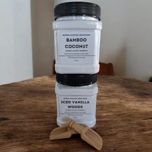 Op de afbeelding: Twee vierkante witte plastic containers met Boros Custom Creations waspoeder, verticaal gestapeld. Het bovenste etiket vermeldt Bamboo Coconut en het onderste etiket Iced Vanilla Woods. Twee kleine houten schepjes liggen ervoor.