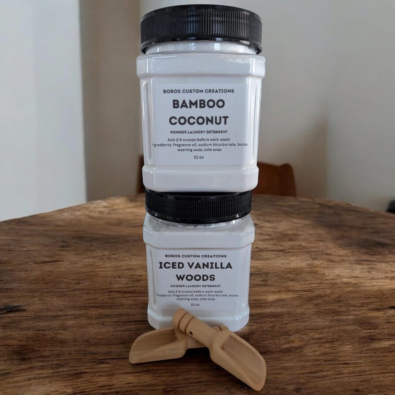 Op de afbeelding: Twee vierkante witte plastic containers met Boros Custom Creations waspoeder, verticaal gestapeld. Het bovenste etiket vermeldt Bamboo Coconut en het onderste etiket Iced Vanilla Woods. Twee kleine houten schepjes liggen ervoor.