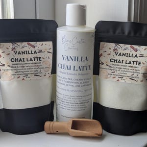 Op de afbeelding: Twee zwart-witte zakjes Vanilla Chai Latte waspoeder, elk gelabeld met 6 oz, naast een witte fles vloeibaar Vanilla Chai Latte wasmiddel en een kleine houten schep voor het afmeten van het waspoeder.