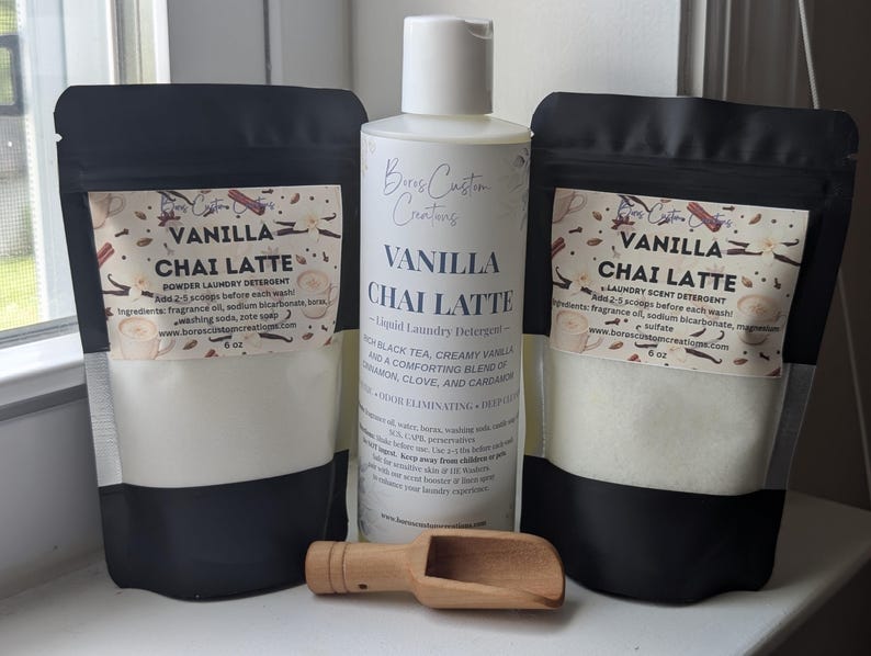 Op de afbeelding: Twee zwart-witte zakjes Vanilla Chai Latte waspoeder, elk gelabeld met 6 oz, naast een witte fles vloeibaar Vanilla Chai Latte wasmiddel en een kleine houten schep voor het afmeten van het waspoeder.