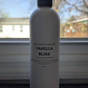 Op de afbeelding: Een witte plastic fles met Boros Custom Creations Vanilla Bliss vloeibaar wasmiddel en een zwarte dop. Het etiket vermeldt ingredi&euml;nten, gebruiksinstructies en het volume van 16 oz. De fles is ondoorzichtig en heeft een cilindrische vorm.