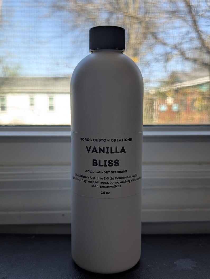 Op de afbeelding: Een witte plastic fles met Boros Custom Creations Vanilla Bliss vloeibaar wasmiddel en een zwarte dop. Het etiket vermeldt ingredi&euml;nten, gebruiksinstructies en het volume van 16 oz. De fles is ondoorzichtig en heeft een cilindrische vorm.