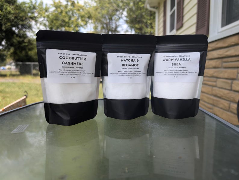Op de afbeelding: Drie zwarte stazakken met wasparfum-booster, elk voorzien van een wit etiket. De etiketten vermelden Boros Custom Creations, met de geuren Cocobutter Cashmere, Matcha & Bergamot en Warm Vanilla Shea, elk aangegeven als 5 oz.