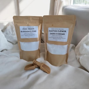 Op de afbeelding: Twee bruine kraftpapieren zakken met waspoeder en witte etiketten. E&eacute;n etiket vermeldt Pink Peach Blossom & Pine, de andere Cactus Flower Jade & Cedar. Elke zak bevat 510 gram product. Een kleine houten schep ligt voor de zakken op de ondergrond.