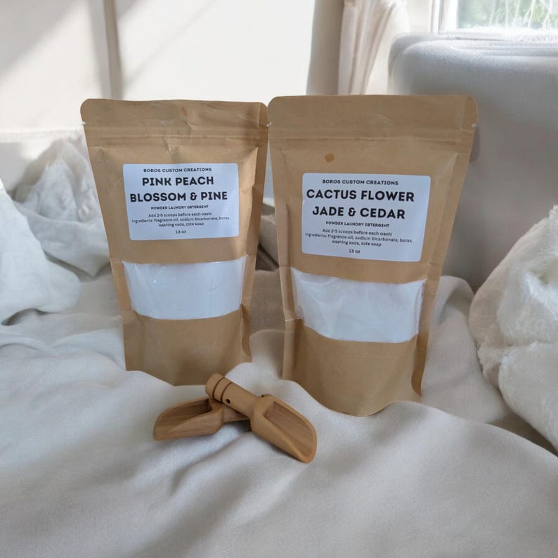 Op de afbeelding: Twee bruine kraftpapieren zakken met waspoeder en witte etiketten. E&eacute;n etiket vermeldt Pink Peach Blossom & Pine, de andere Cactus Flower Jade & Cedar. Elke zak bevat 510 gram product. Een kleine houten schep ligt voor de zakken op de ondergrond.