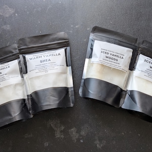 May include: Four black pouches of laundry scent boosters, each with a white label. Two pouches are labelled "Warm Vanilla Shea," and the other two are labelled "Iced Vanilla Woods." The pouches contain a white, granular substance.