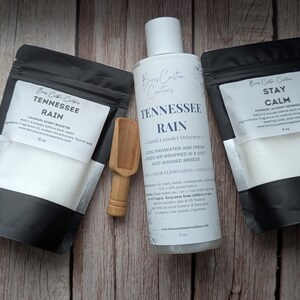 Op de afbeelding: Twee zwart-witte zakjes met het label Tennessee Rain en Stay Calm, een witte fles Tennessee Rain vloeibaar wasmiddel en een klein houten schepje. Elk zakje bevat 227g product en de fles bevat 237ml vloeibaar wasmiddel.