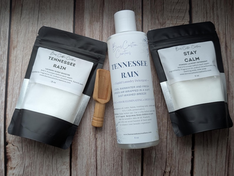 Op de afbeelding: Twee zwart-witte zakjes met het label Tennessee Rain en Stay Calm, een witte fles Tennessee Rain vloeibaar wasmiddel en een klein houten schepje. Elk zakje bevat 227g product en de fles bevat 237ml vloeibaar wasmiddel.