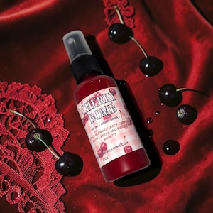 May include: A 2 oz spray bottle with a red liquid and a label reading CHERRY BOMB ROOM + LINEN SPRAY. The label features a pink and white checkered pattern, cherry illustrations, and text describing a blend of juicy cherry, zesty lemon, and merlot.