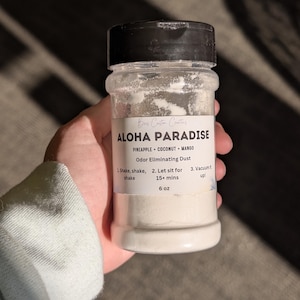 May include: A clear plastic bottle with a black screw-top lid containing white powder. The label reads ALOHA PARADISE, PINEAPPLE COCONUT MANGO, Odor Eliminating Dust, 6 oz, and instructions: 1. Shake, shake, shake, 2. Let sit for 15+ mins, 3. Vacuum it up!