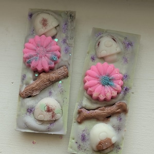 May include: Two rectangular wax melts with a light green and white base, decorated with pink flowers, mushroom shapes, and a brown twig. The melts are sprinkled with purple glitter and have a whimsical, nature-inspired design.