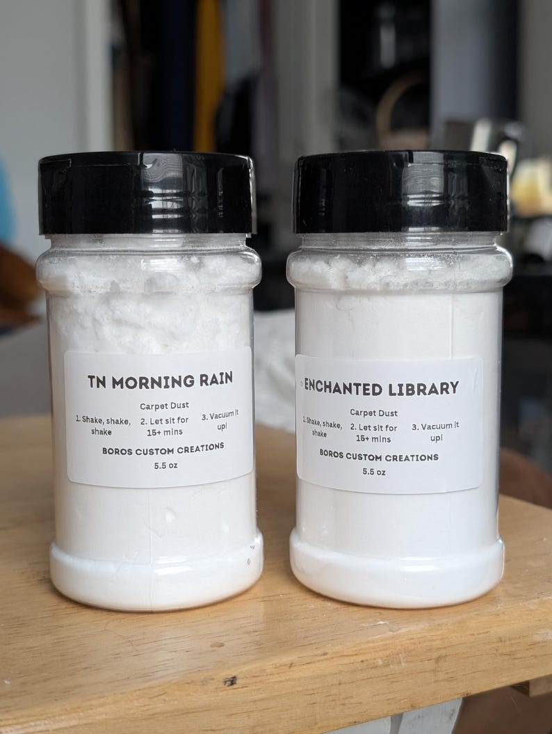 May include: Two clear plastic bottles with black lids containing white carpet dust powder. The labels read TN MORNING RAIN and ENCHANTED LIBRARY, both by BOROS CUSTOM CREATIONS, 5.5 oz, with instructions to shake, let sit for 15+ mins, and vacuum it up.