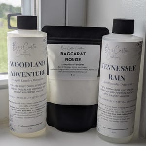 Op de afbeelding: Twee doorzichtige plastic flessen met zwarte doppen gevuld met vloeibaar wasmiddel met de labels Woodland Adventure en Tennessee Rain, naast een zwarte zak met Baccarat Rouge wasparfum, elk voorzien van witte etiketten met zwarte tekst.
